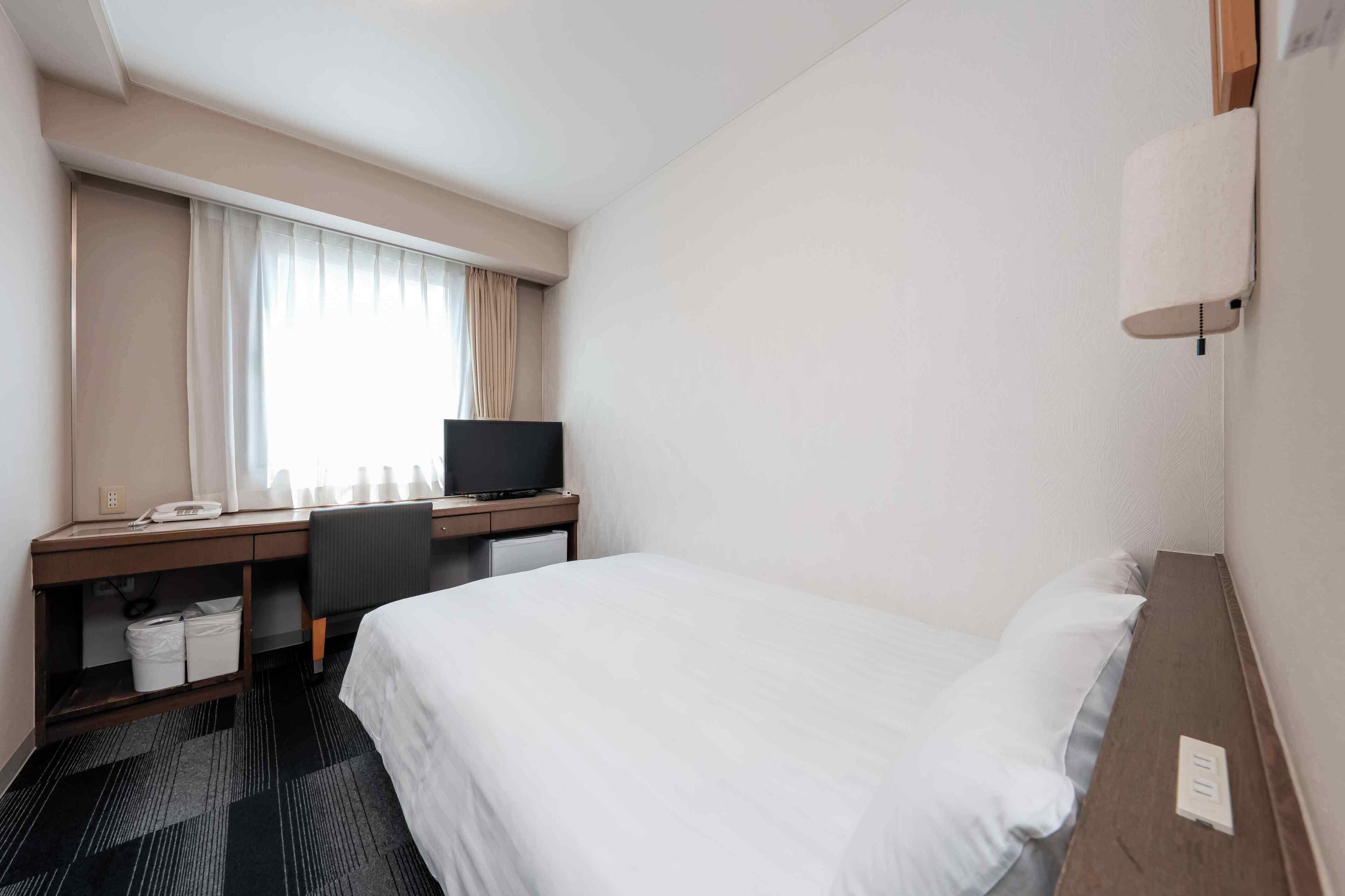 Habitación de hotel con cama, escritorio con computadora, silla, televisor y ventana con cortinas.