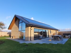 Une maison moderne avec un toit en pente, de grandes fenêtres et un patio aménagé avec du mobilier d’extérieur.