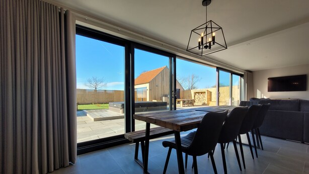 A modern dining area with a large table and chairs, a kitchen area with a TV, and a view of the backyard. - Eastgate Barn, Ashlin Farm Barns (Lincoln)