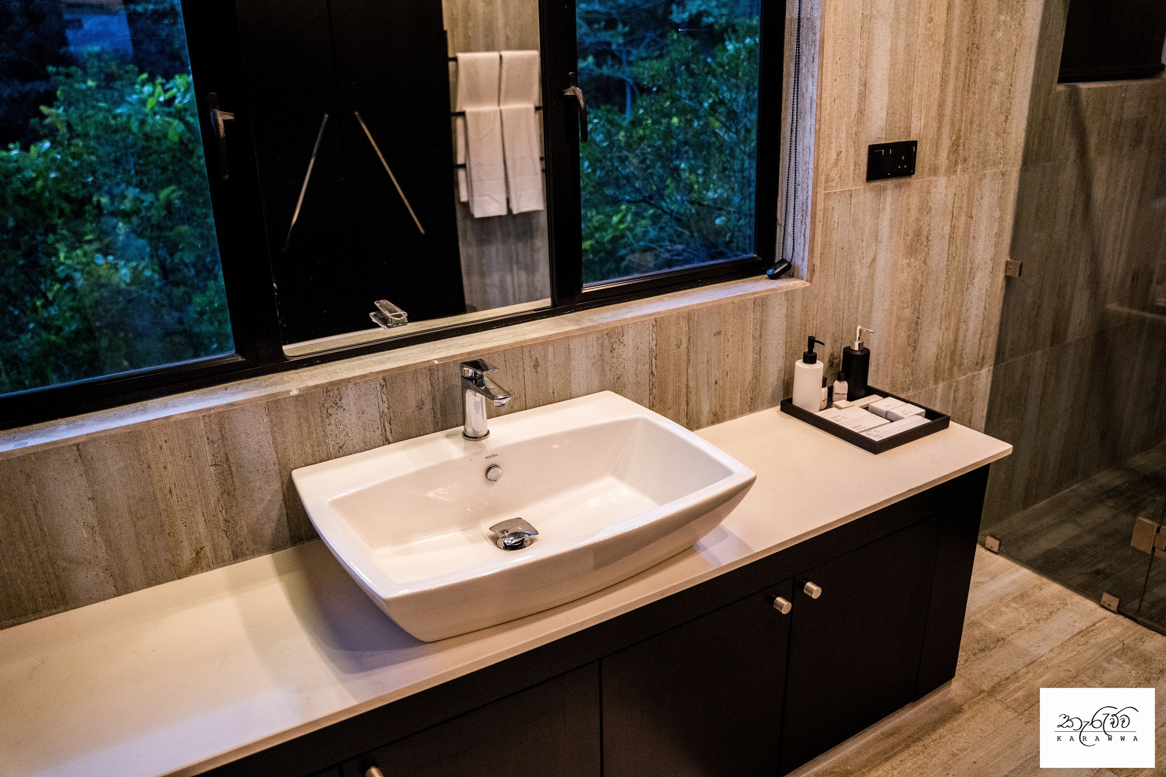 A modern bathroom with a large sink, a mirror, and a window with a view of greenery.