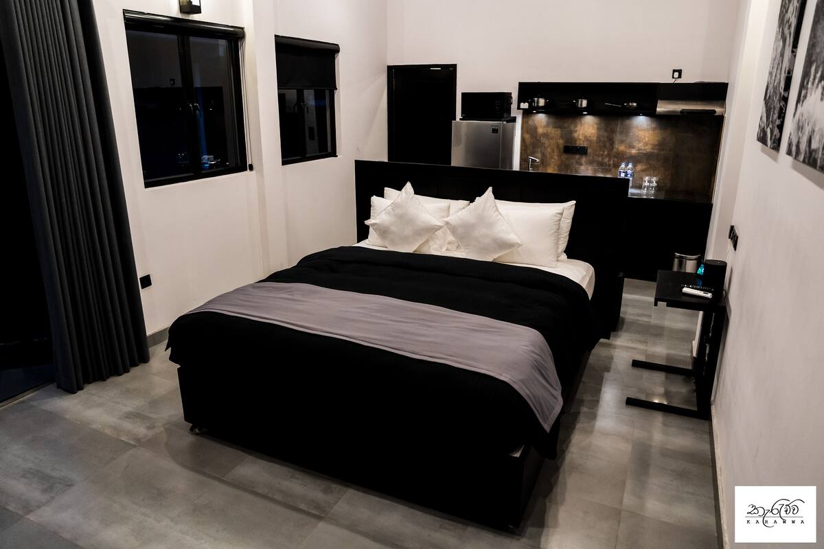 A modern bedroom with a large bed, a small bedside table, a built-in wardrobe, and a kitchen area in the background.