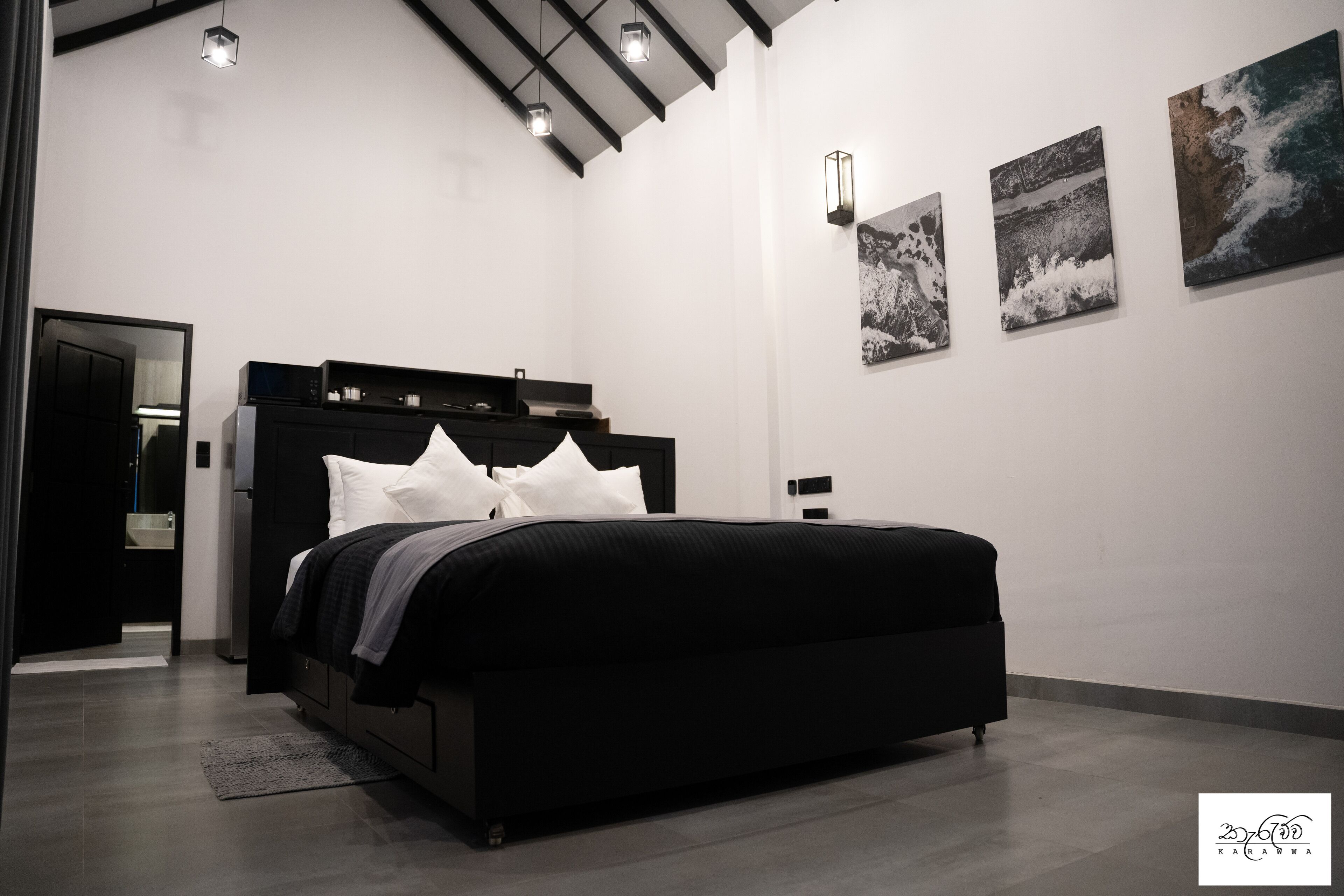 A modern bedroom with a black bed, white pillows, and a wooden headboard. There are three wall-mounted artworks depicting abstract and natural scenes.