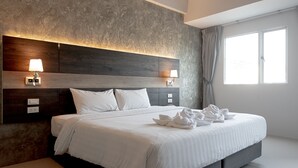 A modern hotel room with a large bed, two bedside lamps, a headboard with a textured design, and a view of the outside through a window. - The Proud Exclusive Hotel (Nakhon Pathom)