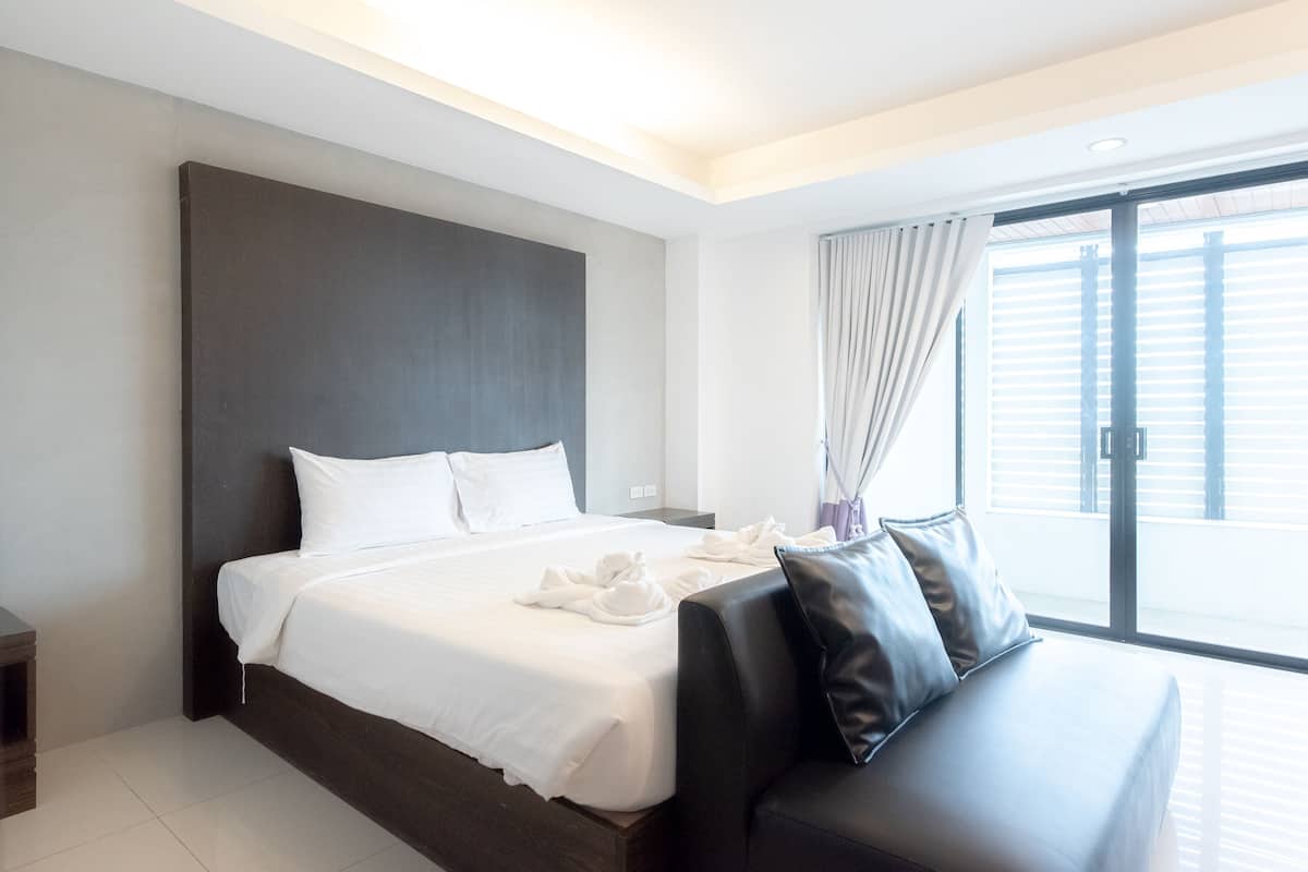 A modern hotel room with a large bed, a dark headboard, a black leather sofa, and a large window with blinds.