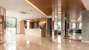 A modern hotel lobby with a reception desk, wooden accents, and large windows offering views of the outdoors. - The Proud Exclusive Hotel (Nakhon Pathom)