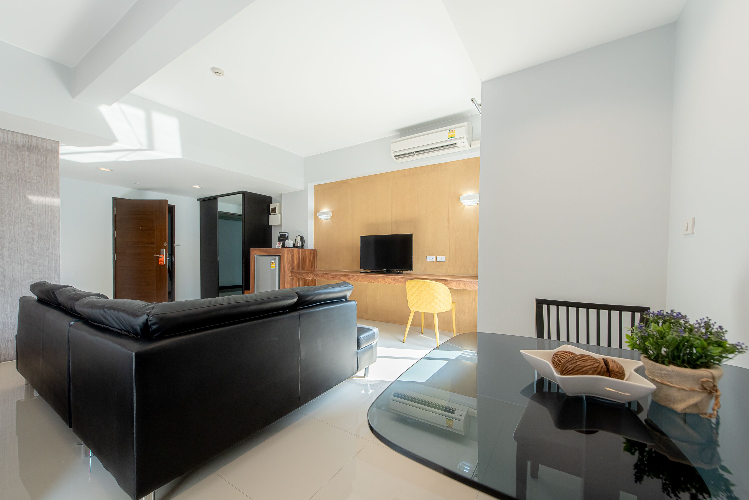 A modern living room with a black sofa, a glass dining table, and a wooden kitchen area in the background.