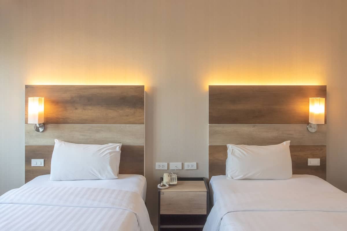 A hotel room with two beds, each with a white pillow and a headboard with a light fixture. There is a nightstand between the beds with a phone and a lamp.
