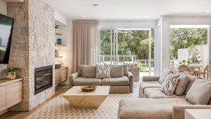 Un salon moderne avec une cheminée en pierre, un ensemble de canapés beige, une table basse et une grande fenêtre donnant sur l’extérieur.