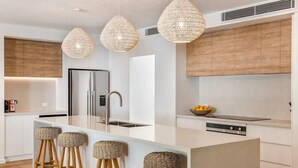 Une cuisine moderne dotée d’un îlot central, d’appareils électroménagers en acier inoxydable et de trois luminaires à abat-jour en fibres tressées.