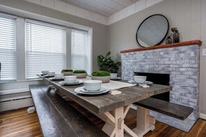 A dining area with a long wooden table set for four, a brick fireplace, and a large window with blinds.