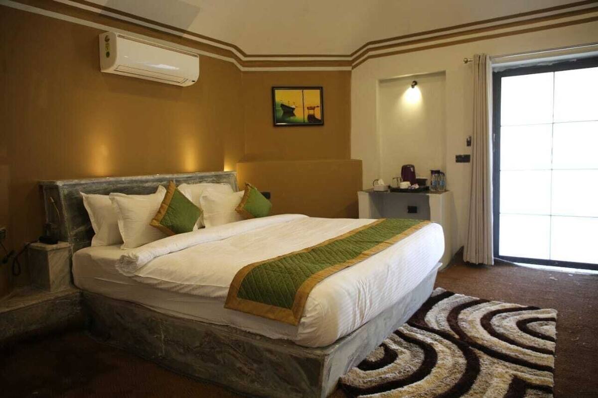 A hotel room with a bed, a nightstand, a wall-mounted air conditioner, a painting, and a view through a door.