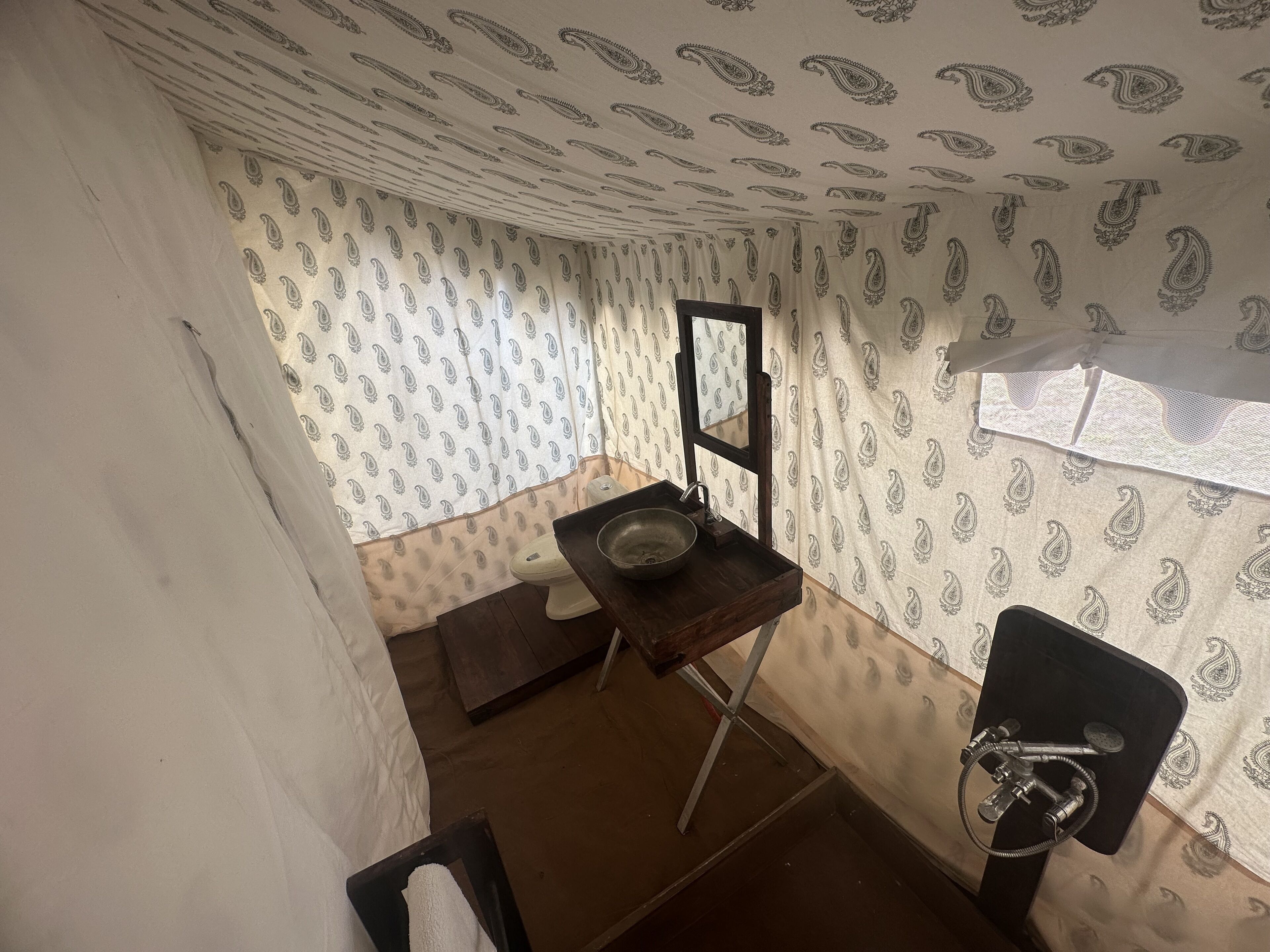Et lille, kompakt badeværelse med toilet, vask og bruseområde. Væggene er dækket af tapet med paisley-mønster.