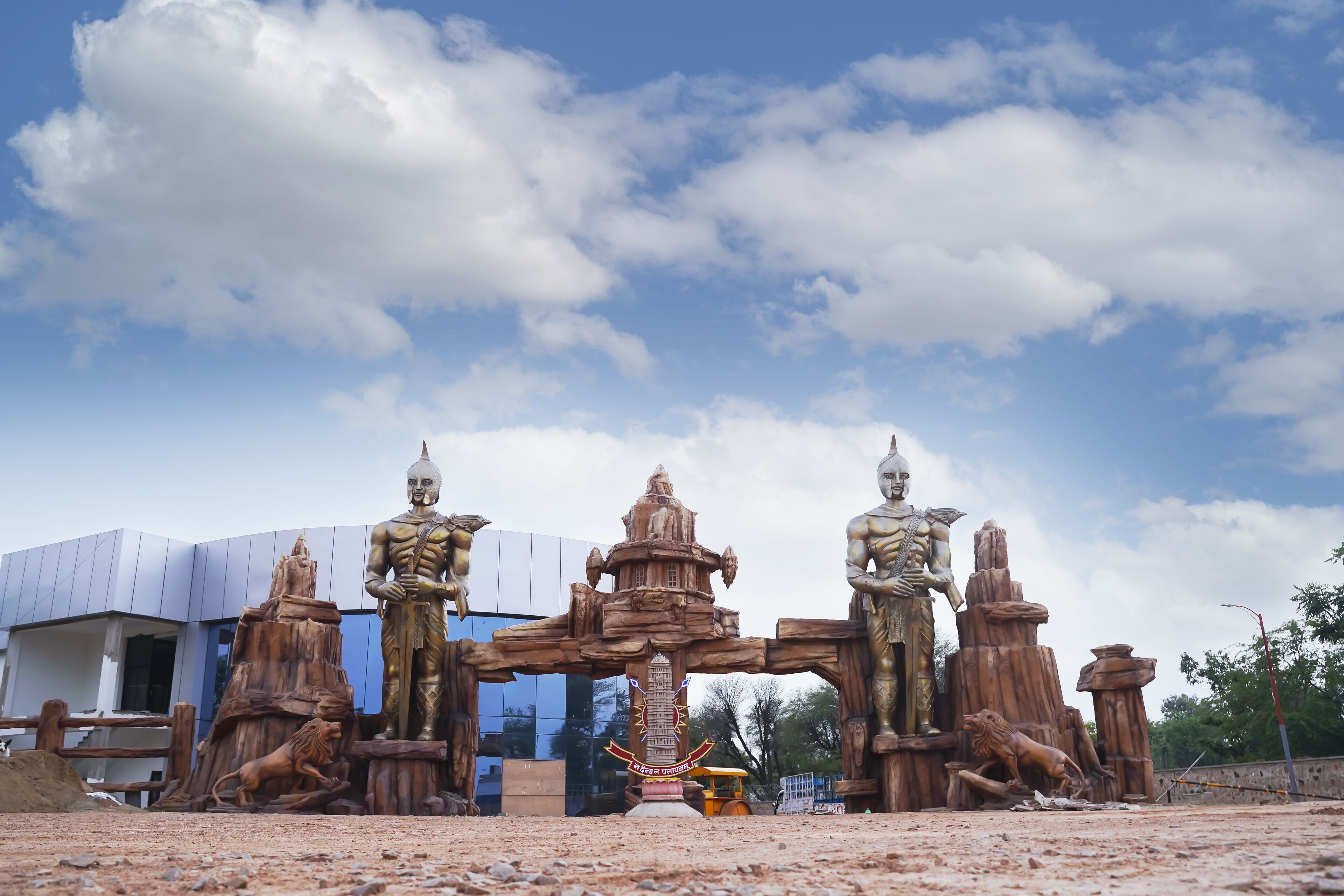 A large outdoor sculpture with multiple statues and a central monument, set against a modern building and a clear sky.