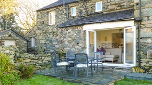 A stone house with a patio area featuring a table and chairs, a sliding glass door leading to a kitchen, and a stone wall surrounding the property. - Riverside Cottage  - Duddon Valley (Dog-friendly) (Seathwaite)