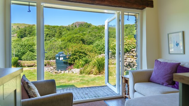 A room with a large sliding glass door leading to a garden with a water feature and a view of a hill. - Riverside Cottage  - Duddon Valley (Dog-friendly) (Seathwaite)