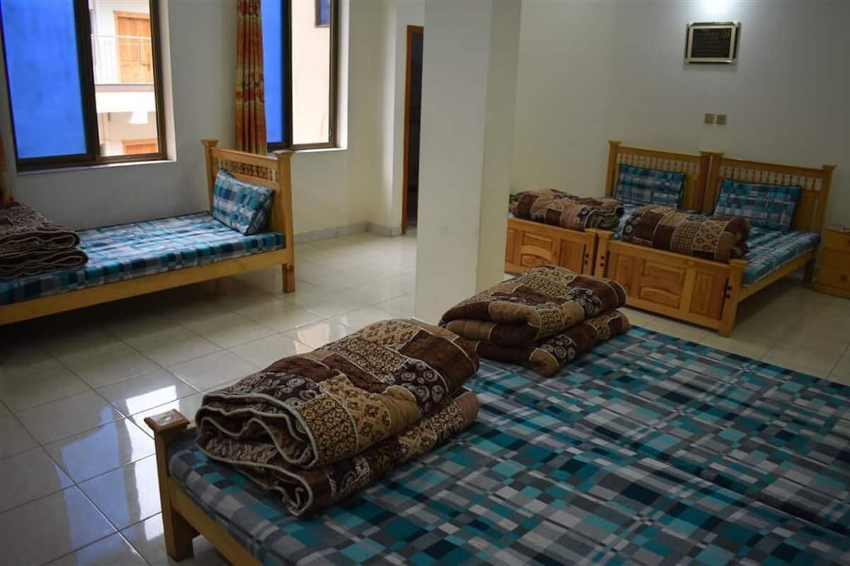 A room with two single beds and a larger bed, all with patterned bedding. There are windows with curtains, a tiled floor, and a wall-mounted air conditioning unit.