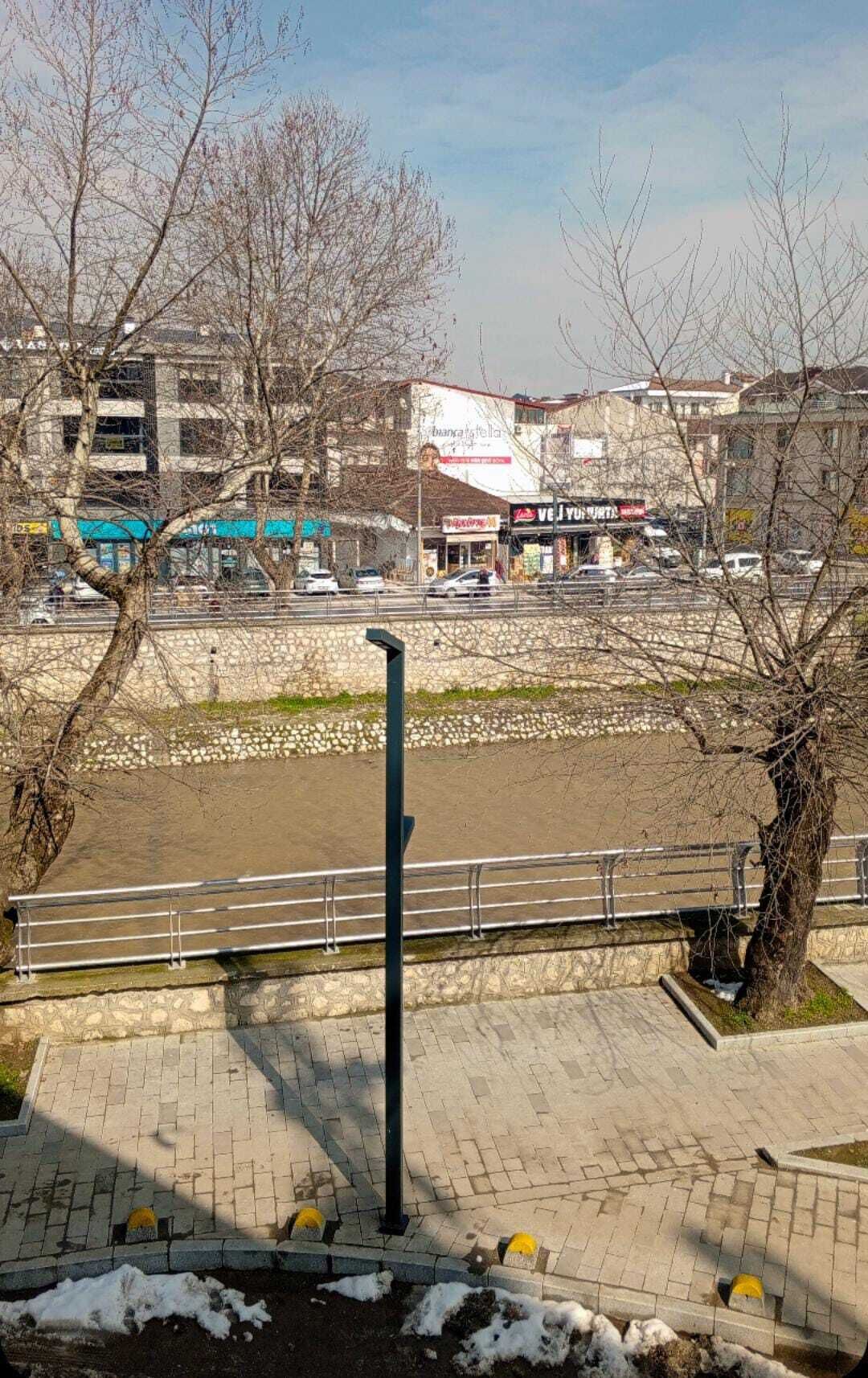 Uma passarela pavimentada com guarda-corrimão de metal, um muro de pedra e um rio ao fundo.