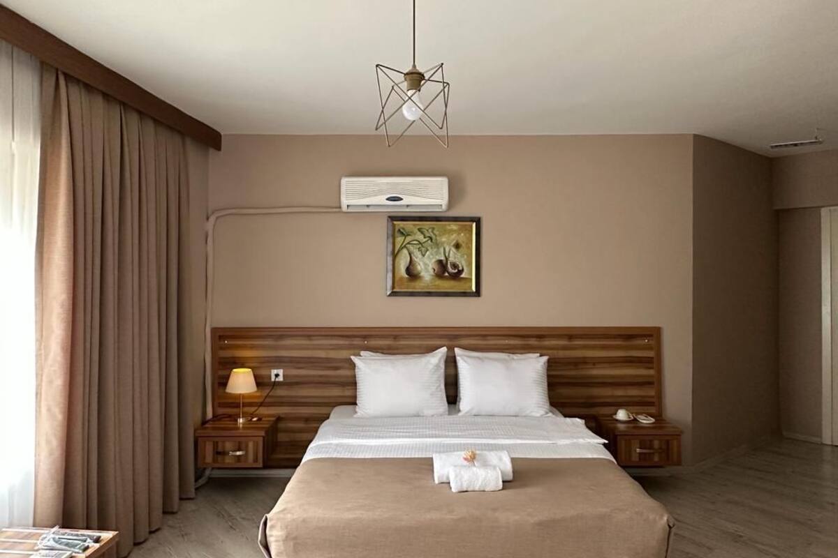 A hotel room with a large bed, a wooden headboard, a bedside table with a lamp, a wall-mounted air conditioner, and a framed artwork above the bed.