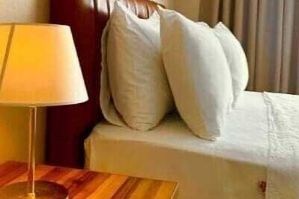 A hotel room with a wooden side table, a lamp, a bed with white bedding, and a wall with decorative patterns.