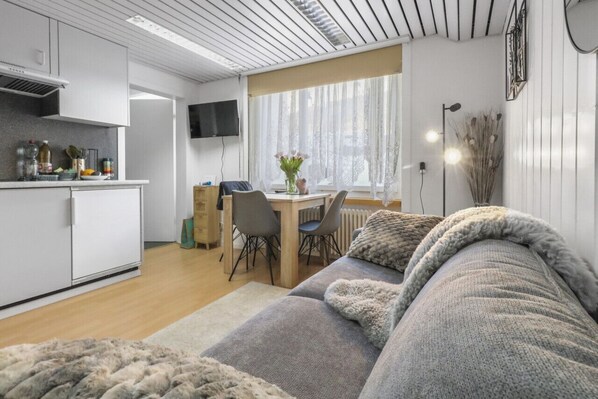 A modern, compact living space with a kitchenette, a dining area, and a cozy seating area with a sofa and a large window providing natural light.