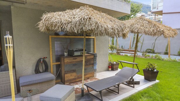 A modern outdoor patio with two thatched umbrellas, a wooden cabinet, a lounge chair, and a small table. - Nice apartment near the center (Martigny)