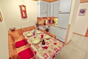 A compact kitchen with white cabinets, a stove, and a refrigerator. A dining table with red cushions and a patterned tablecloth is set up. - Excellent apartment with free covered parking (Lens)