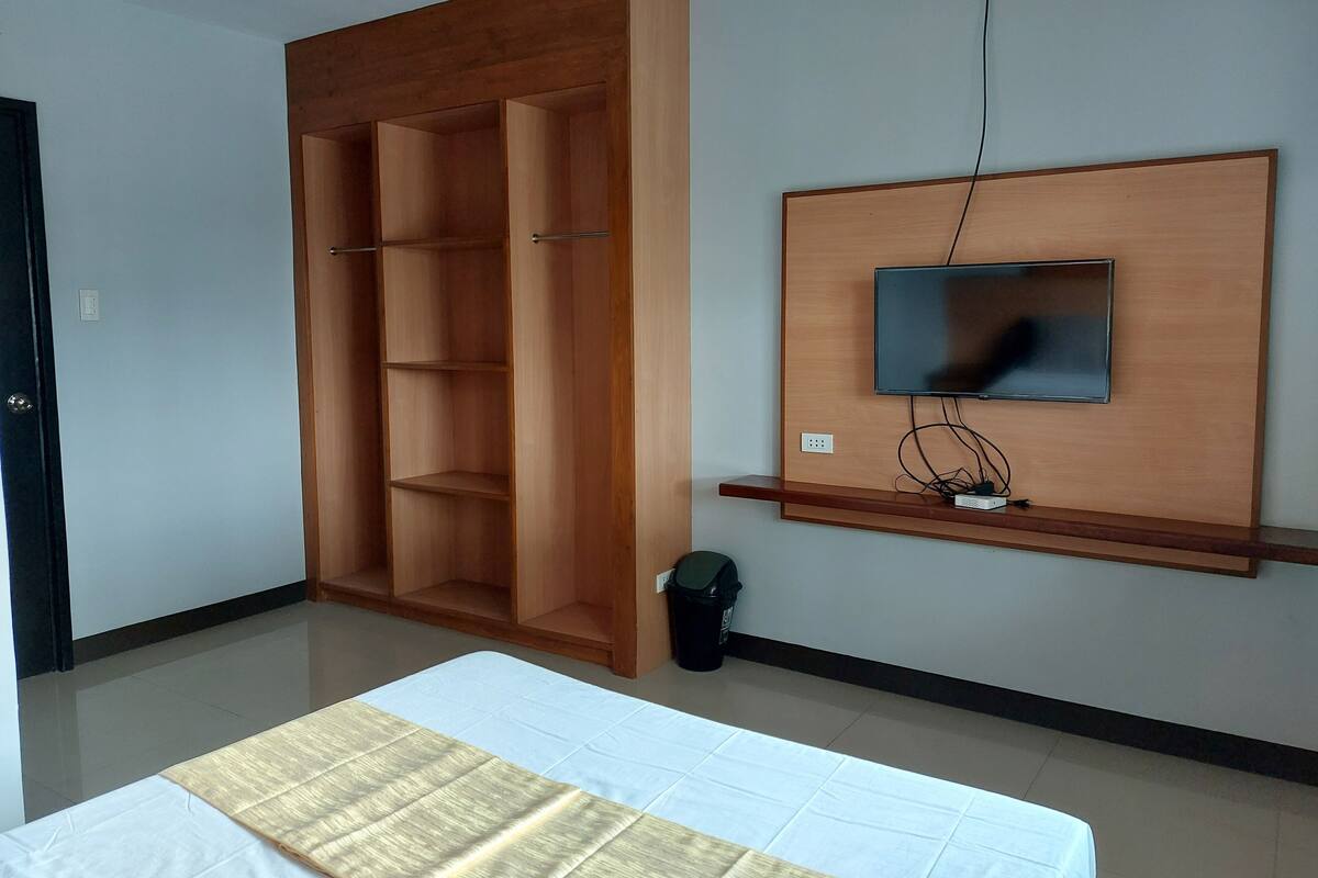 Ein Schlafzimmer mit einem Bett, einem Holzschrank, einem an der Wand befestigten Fernseher und einem Mülleimer.