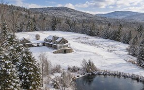 Une grande maison avec des fondations en pierre et une véranda qui fait le tour de la maison, entourée d’arbres couverts de neige et d’un lac gelé.