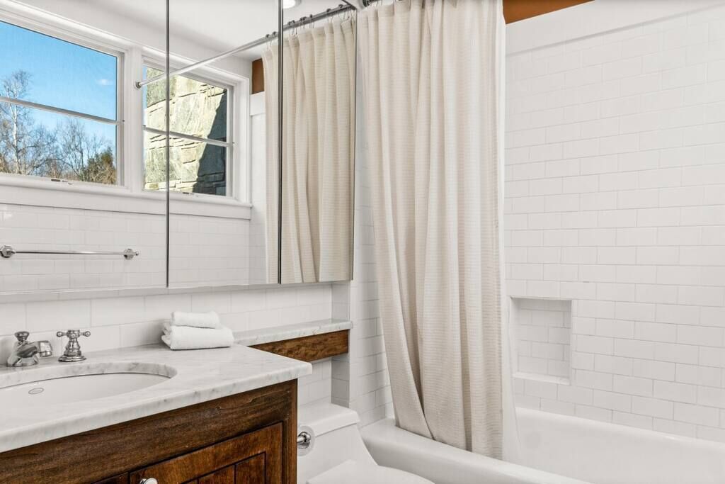 A bathroom with a white sink, wooden vanity, a mirror, a bathtub, and a window with a view of trees.