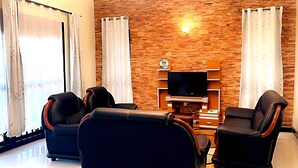 A living room with two black leather sofas, a flat-screen TV on a wooden wall, and a small wooden cabinet. - Cosy Family Home (Kampala)