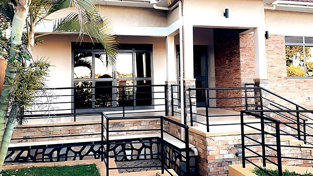 A modern two-story house with a brick exterior, a balcony with a metal railing, and a palm tree in the foreground. - Cosy Family Home (Kampala)