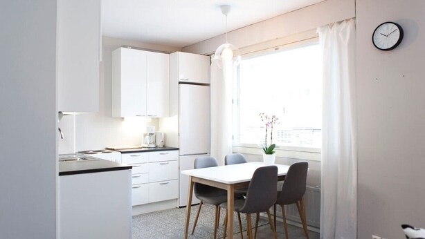 Una cocina moderna con gabinetes blancos, una mesa de comedor con sillas, un reloj en la pared y una ventana con cortinas.