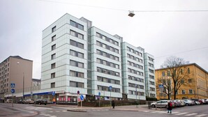 Un edificio de varios pisos con fachada moderna de vidrio, un paso de peatones y un área de estacionamiento con automóviles.