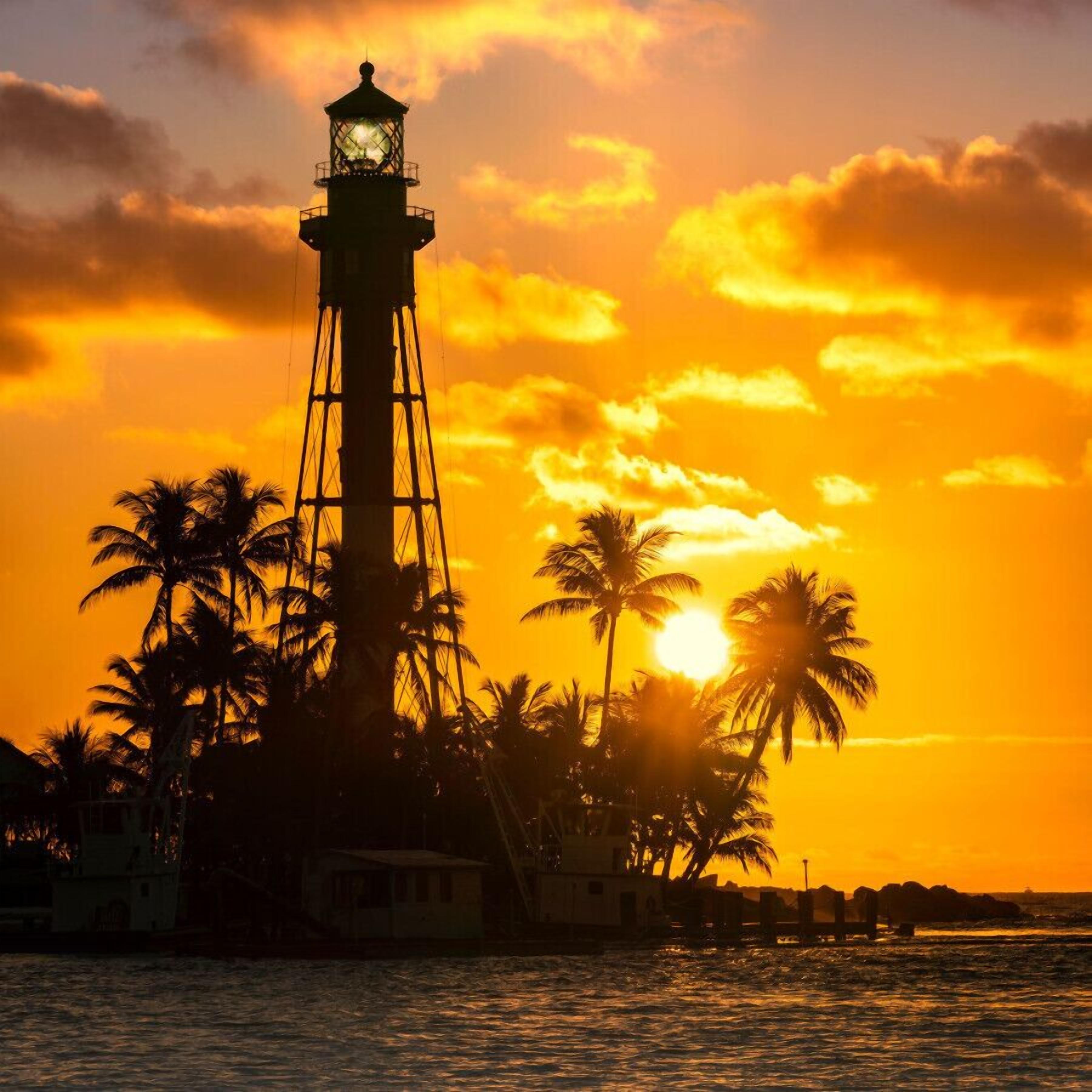 Un phare avec une lanterne au sommet, entouré de palmiers et avec un coucher de soleil en arrière-plan.