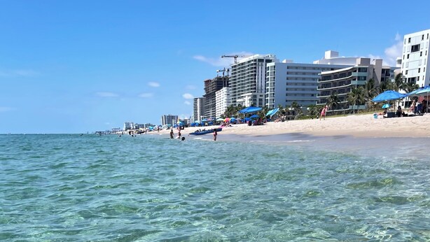 A beach with clear water, buildings, and umbrellas.