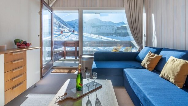 Une pièce moderne avec un canapé bleu, un meuble en bois et une table en verre sur laquelle se trouvent une bouteille et des verres. Un balcon avec vue sur un paysage enneigé et des bâtiments est visible à travers de grandes fenêtres.