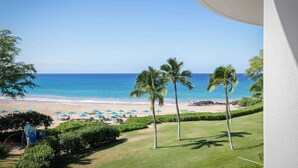 Vista a la playa desde un balcón con palmeras, arena y sombrillas.