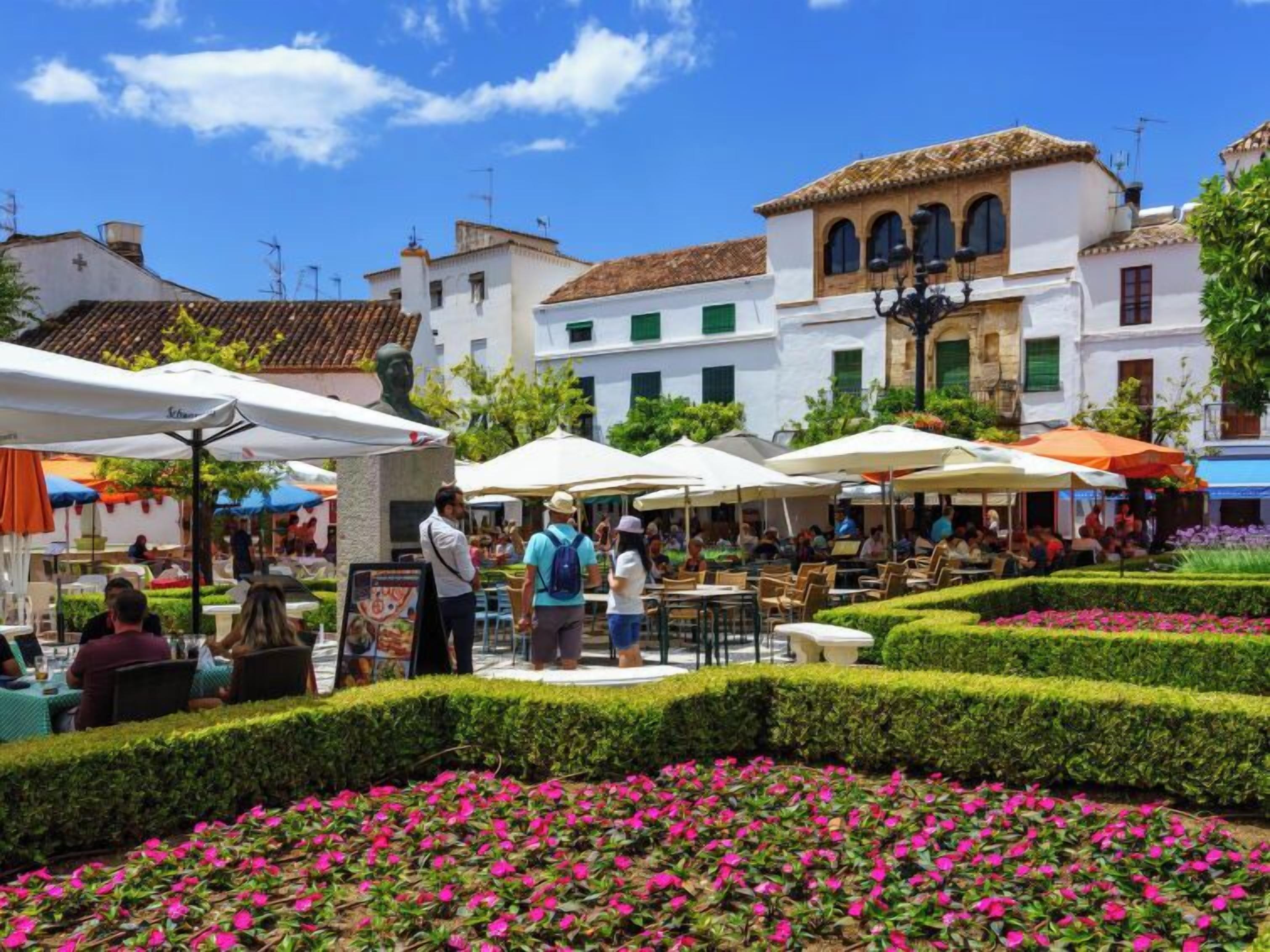 Un café en plein air, avec des tables et des chaises, entouré de haies soigneusement taillées et de parterres de fleurs aux couleurs vives.