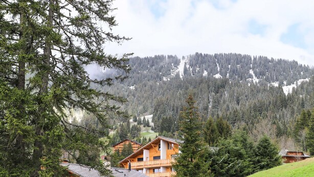 En træchalet med balkon, omgivet af et skovklædt bjerglandskab.
