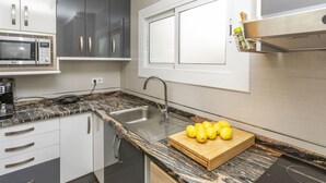 A modern kitchen with a granite countertop, stainless steel sink, and a microwave. There are cabinets with silver handles and a wooden cutting board with lemons on the counter.