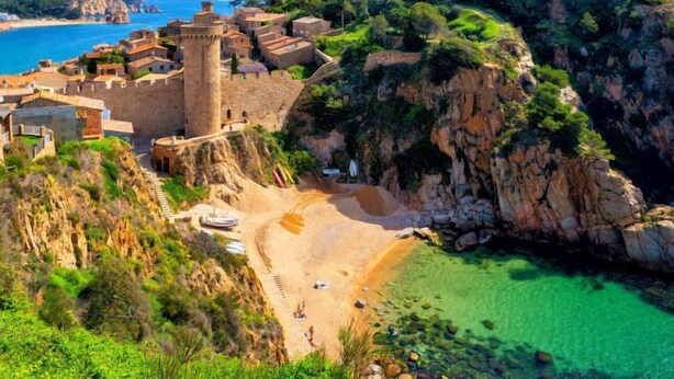 A coastal town with a historic stone castle, a sandy beach, and clear turquoise waters.