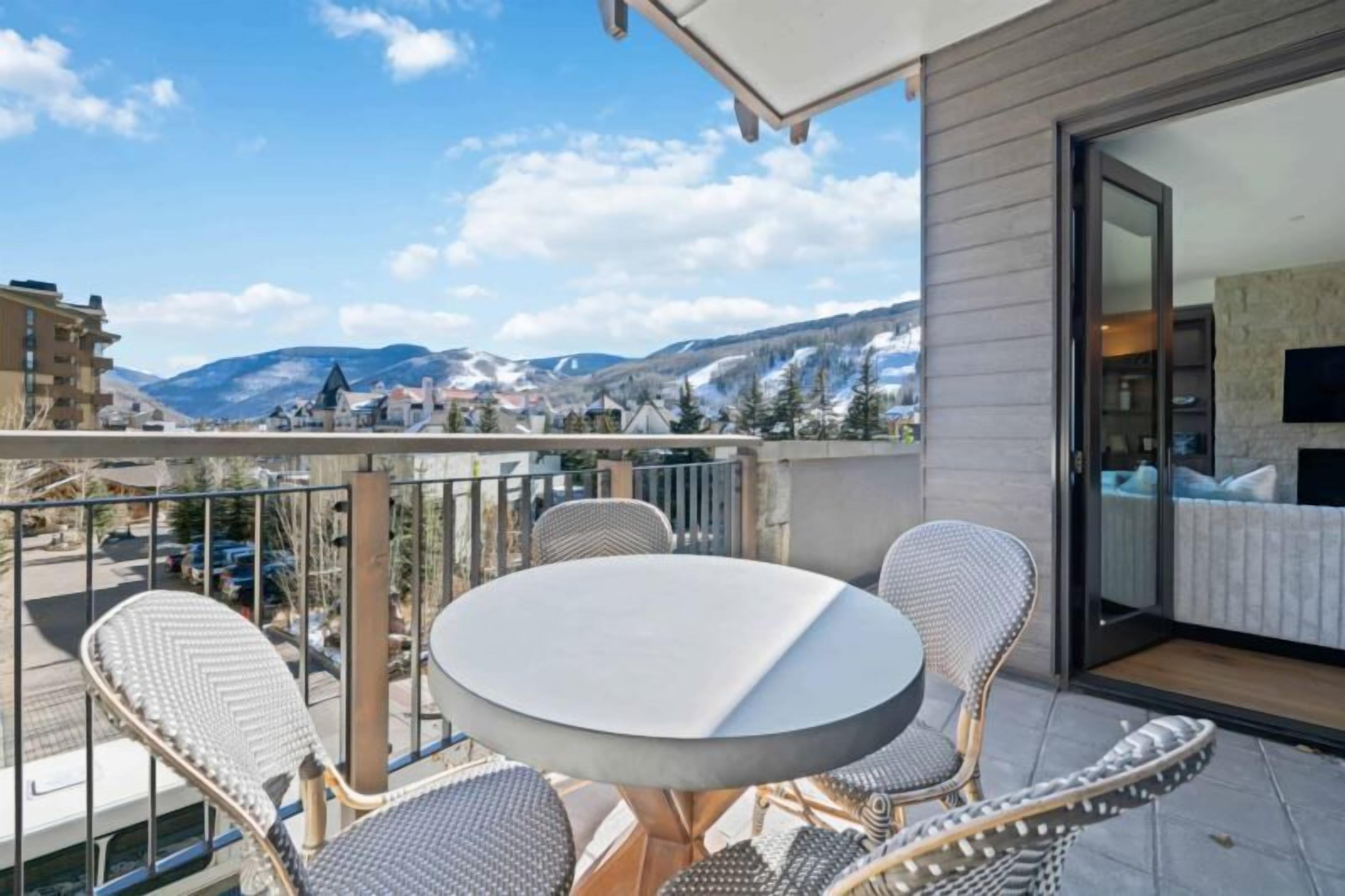 Un balcón con una mesa redonda y sillas, una puerta de vidrio que da a una habitación, y vistas a la montaña.