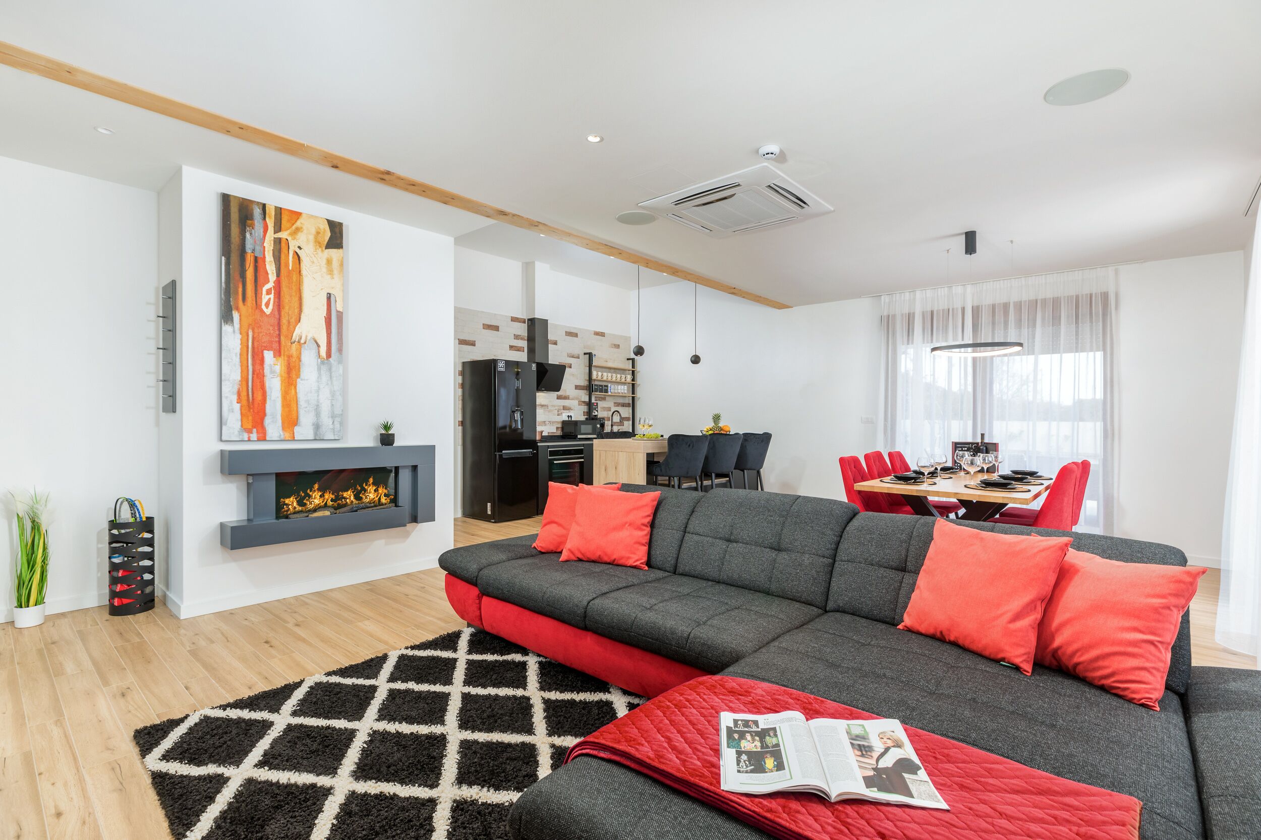 A modern living room with a grey sofa, red cushions, a black and white rug, a fireplace, and a dining area in the background.