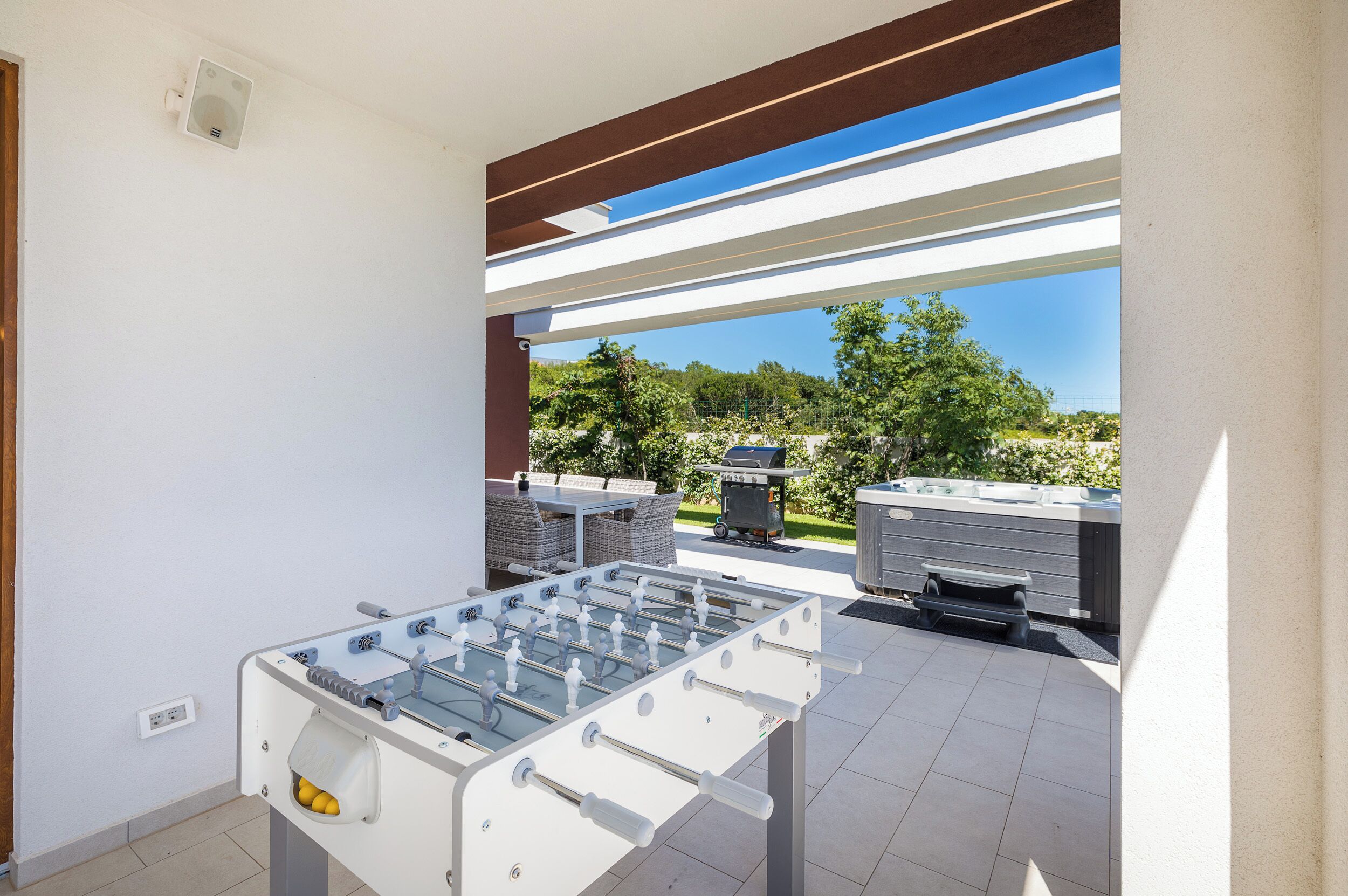 A foosball table in a covered outdoor area with a view of a barbecue grill and outdoor seating.