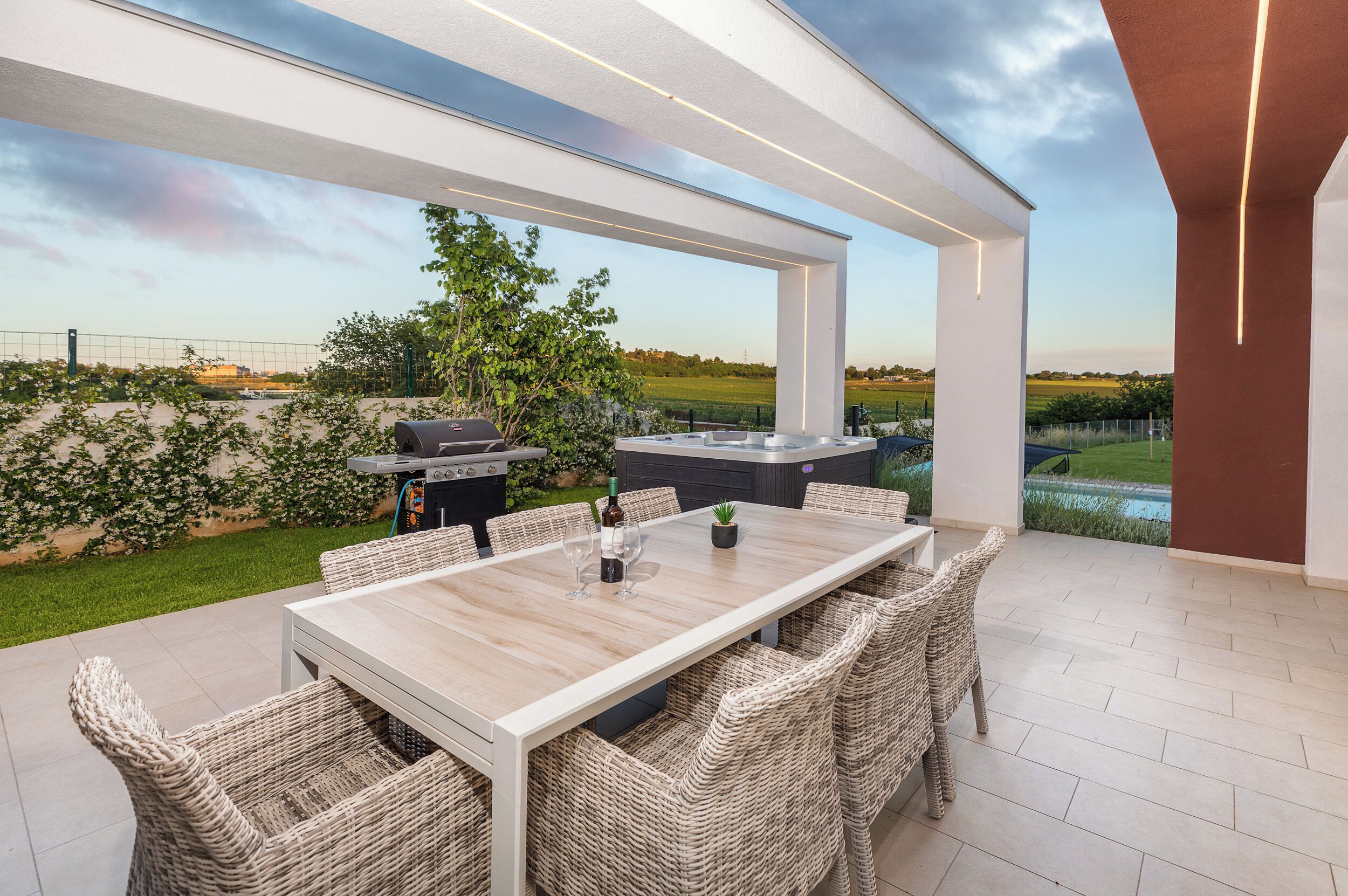Patio esterno con tavolo e sedie, un barbecue e vista su un campo e il cielo.