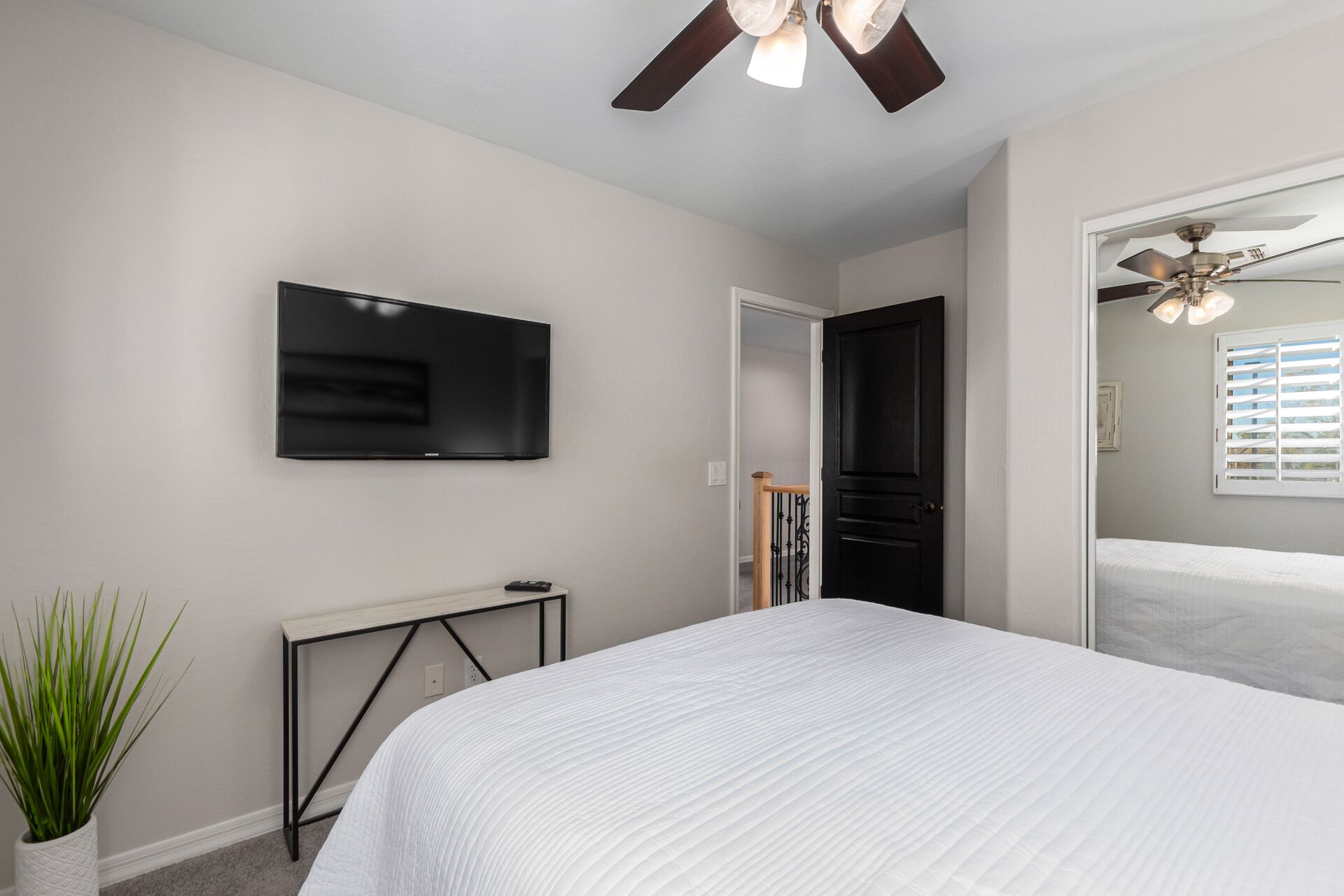 A bedroom with a bed, a TV mounted on the wall, a small table, and a potted plant.