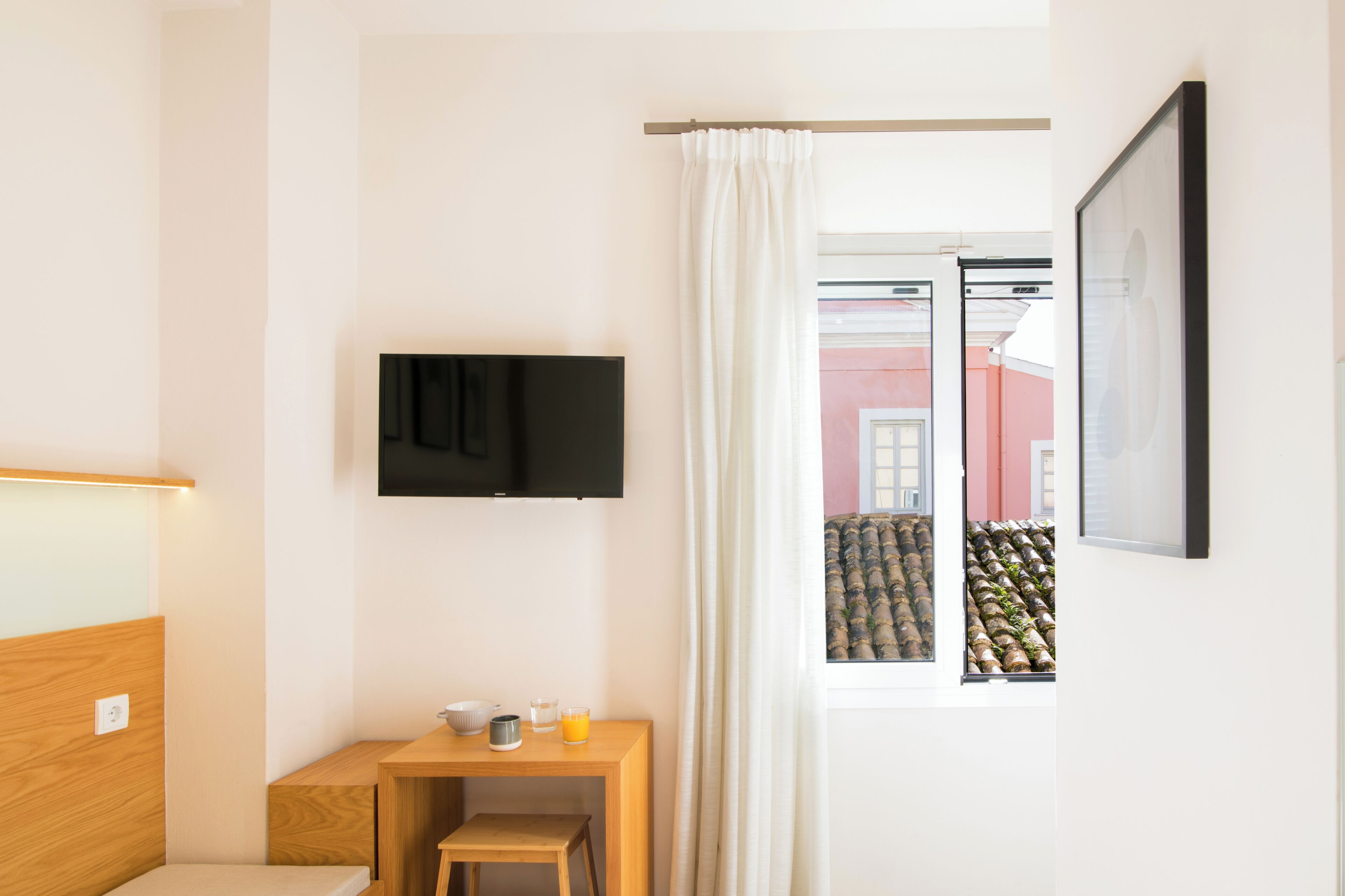 Et moderne badeværelse med en træbænk, et lille bord med toiletartikler, et tv ophængt på væggen, og et vindue med udsigt til en bygning med stablede fade udenfor.