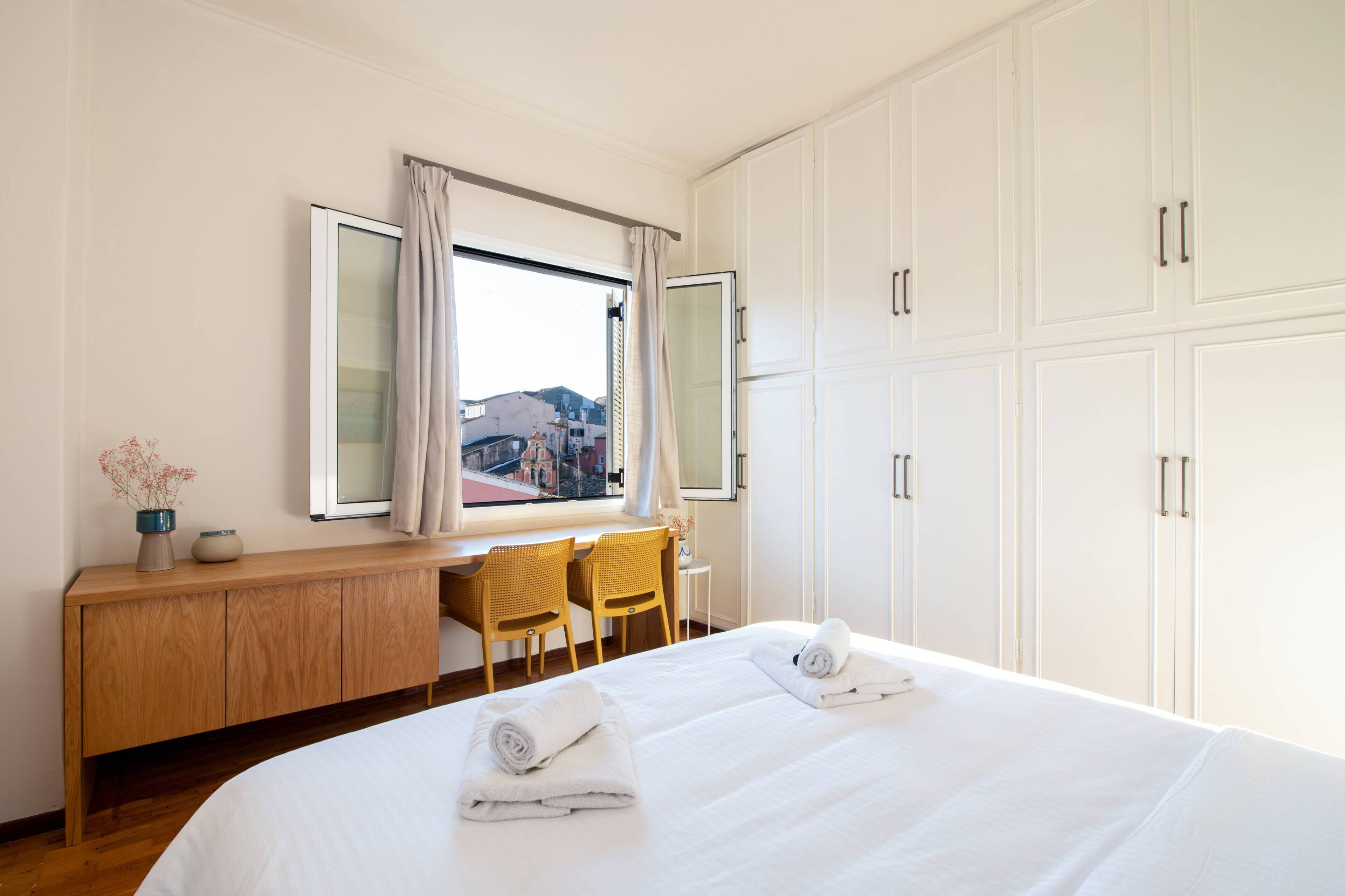 A hotel room with a large bed, a wooden desk, two chairs, a window with curtains, and a view of buildings outside.