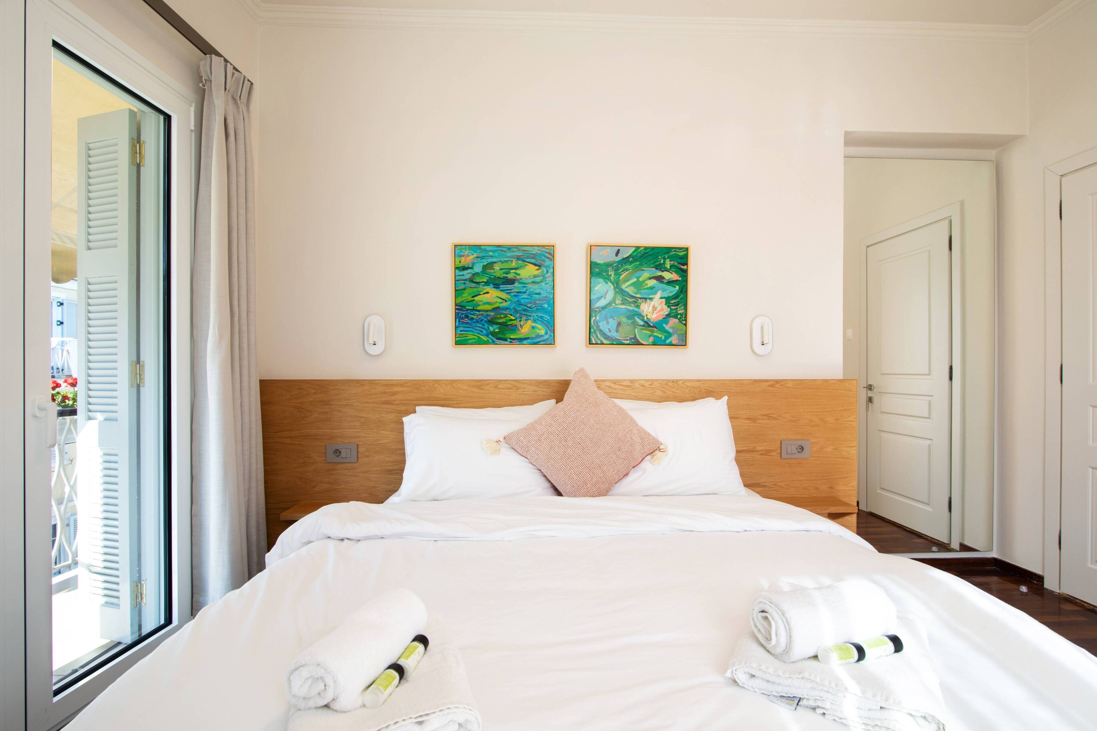 A hotel room with a bed, two paintings on the wall, a window with curtains, and a door.
