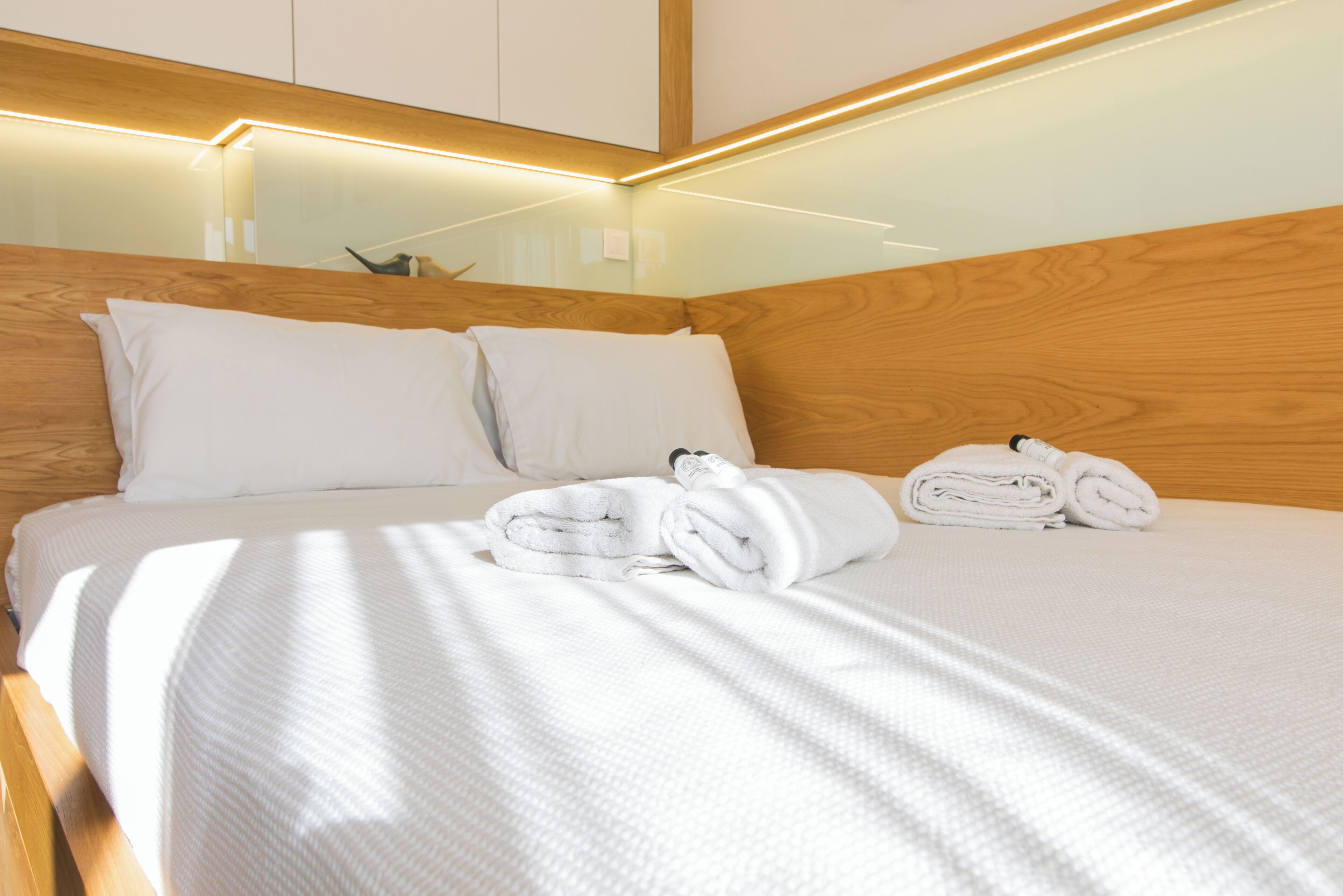 Una cama bien hecha con ropa de cama y toallas blancas, cabecera de madera e iluminación empotrada.